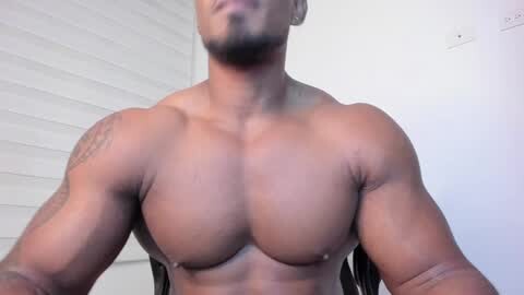 Media: Video of a muscular, shirtless Black man with a trimmed beard, showcasing his broad shoulders, defined chest, and arms. Background features a white wall with a slight texture and a power outlet.