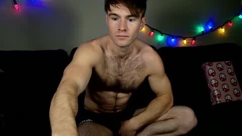 Media: Video of a young, fit, shirtless man with short brown hair, sitting on a black couch, wearing black underwear, surrounded by colorful string lights, with a pink pillow and gift wrap in the background.
