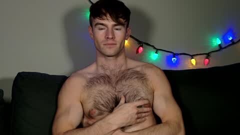 Media: Video of a shirtless, muscular, fair-skinned man with short brown hair, wearing a Christmas sweater with a large chest of dark hair, sitting on a dark couch, illuminated by colorful string lights in the background.