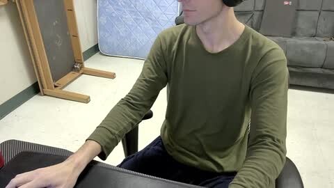 Media: Video of a slim, fair-skinned man with short dark hair in a green long-sleeve shirt, sitting at a black desk in a room with beige walls and a blue quilted pad.