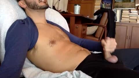 Media: Video of a shirtless, fair-skinned man with a trimmed beard and short hair, lying on a couch, holding a large, erect, circumcised penis. Background includes a wooden desk, office chair, and bookshelves.