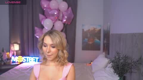Media: Video of a blonde woman in a pink dress holding a bouquet of balloons in a pastel-themed bedroom with a bed, potted plants, and abstract art on the wall.