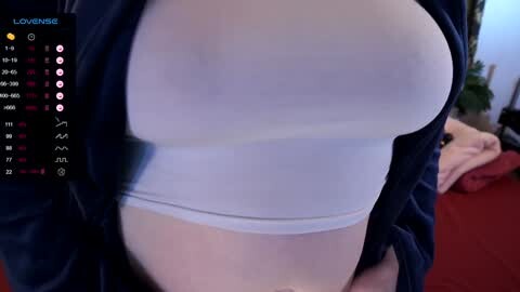 Media: Video of a woman wearing a tight white tank top, revealing her large, round breasts. Background shows a messy room with red carpeting and a calendar displaying \"LOVEBITE\" on the left.