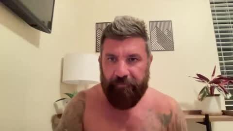 Media: Video of a shirtless man with a full beard and short gray hair, wearing a black tank top, standing in a bedroom with a TV, potted plants, and white blinds.