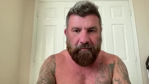 Media: Video of a middle-aged, bearded man with a muscular build and numerous tattoos on his arms, standing shirtless in front of white double doors.