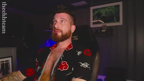 Video of a bearded man with short brown hair, wearing a black Hawaiian shirt with red and white patterns, sitting in a gaming chair, dimly lit room with a TV showing a racing game.