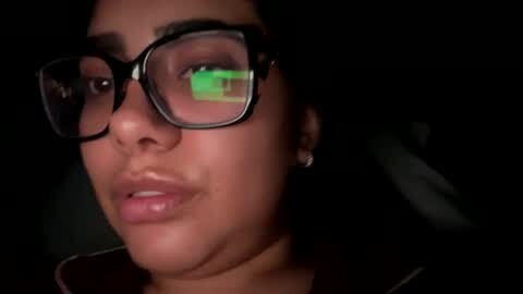 Media: Video of a woman with medium-brown skin, wearing black-framed glasses, short curly hair, and a black jacket, illuminated by a green light, giving a mysterious, otherworldly look.