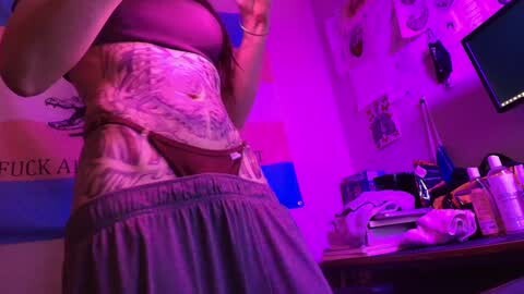 Media: Video of a tattooed woman in a purple bra and grey skirt, standing in a dimly lit room with purple lighting, surrounded by cluttered shelves and posters.