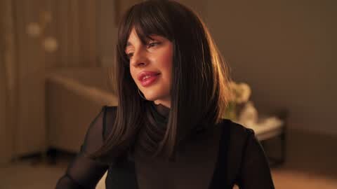 Media: A video of a woman with straight, dark brown hair and fair skin, wearing a sheer black top, smiling in a warmly lit, softly blurred bedroom setting.