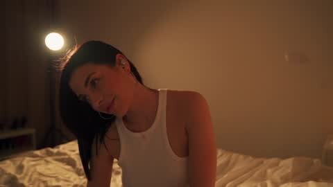Media: Video of a young woman with long, dark hair, wearing a white tank top, sitting on a bed with rumpled white sheets in a dimly lit, cozy bedroom.