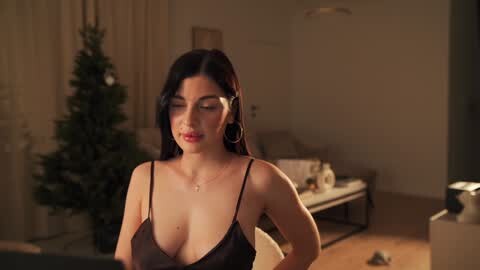 Media: Video of a young woman with long black hair, wearing a black lace bra, standing in a dimly lit living room with a decorated Christmas tree in the background.