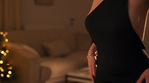 Media: Video of a slender woman in a black, sleeveless dress, showcasing her slim waist and small breasts. She stands in a dimly lit living room with a Christmas tree and a sofa in the background.