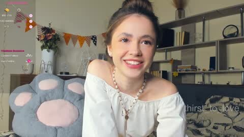 Media: Video of a smiling young woman with light skin, brown hair, and a white off-shoulder top, sitting on a bed in a cozy, modern room with shelves and a floral centerpiece.