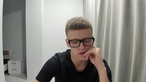 Media: Video of a young man with short, light brown hair, wearing black glasses and a black shirt, resting his head on his hand, seated indoors against a white wall and gray curtain backdrop.