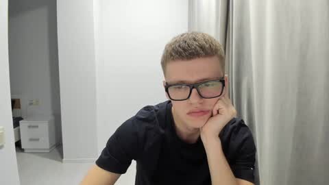 Media: Video of a young man with short, light brown hair and glasses, wearing a black T-shirt, resting his chin on his hand, in a modern, white-tiled room with a metallic curtain and a visible door to a restroom.