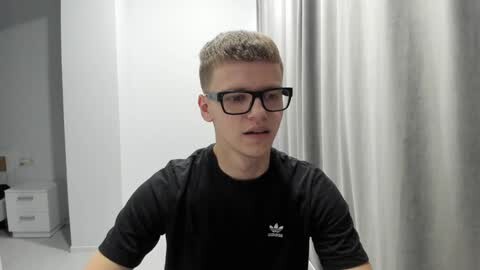 Media: Video of a young, fair-skinned man with short, light brown hair and black-framed glasses, wearing a black Adidas shirt, speaking in a modern, white-walled room with grey curtains.
