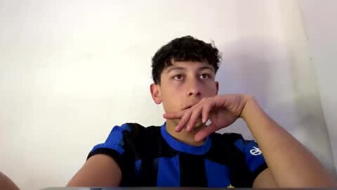 Media: Video of a young boy with curly hair and light skin, wearing a blue and black striped shirt, sitting against a plain white wall, looking thoughtful with his hand on his chin.