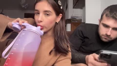 Media: Video of a young woman with dark hair, wearing a beige jacket, drinking from a purple water bottle, and a young man with dark hair, wearing a black shirt, looking at his phone, in a living room.