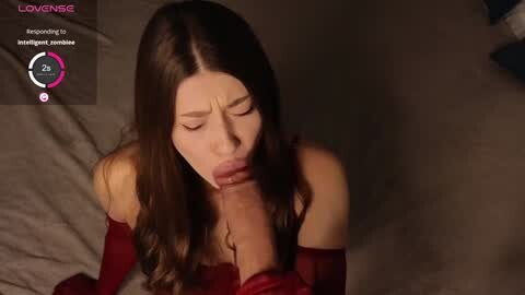 Media: Video of a young woman with long brown hair, wearing a red off-shoulder dress, performing oral sex on a large erect penis. Background shows a bed with gray sheets, dim lighting.