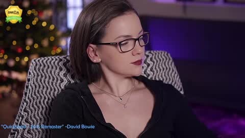 Video of a fair-skinned woman with straight, shoulder-length brown hair, wearing glasses and a black sweater, seated on a patterned couch, with a festive, blurred Christmas tree in the background.