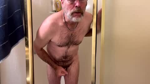 Media: A video of a naked older white man with a full beard and glasses standing in a shower, holding his penis. The background features a beige wall and a blue towel.