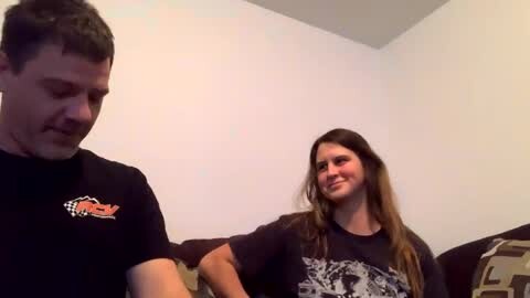 Media: Video of a young couple sitting on a couch, man in black t-shirt with a checkered logo, woman in black graphic t-shirt, smiling. Background is a plain, light-colored wall.