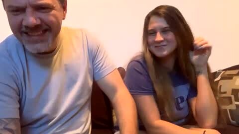 Media: Video of a middle-aged man with a mustache and beard, wearing a light blue T-shirt, and a young woman with long, straight, light brown hair, wearing a purple T-shirt, seated on a patterned couch against a plain wall.