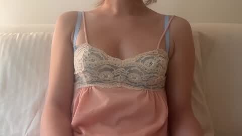 Media: Video of a light-skinned woman with a slim build, wearing a delicate lace-trimmed, pastel pink satin camisole. The background is a plain, off-white wall and a white, textured pillow.