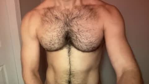 Media: Video of a light-skinned, muscular man with a full chest of dark, curly body hair, standing in a plain room with a white wall and door in the background.