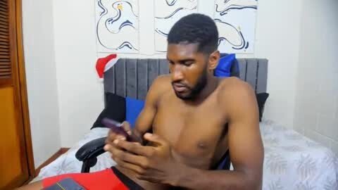 Media: Video of a shirtless, muscular Black man with short hair, seated on a bed with gray cushions, wearing red and blue pants, playing with a smartphone in a simple bedroom with white walls and abstract art.