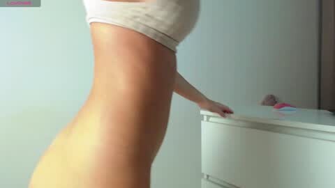 Media: Video of a slender, fair-skinned woman in a tight, white crop top, standing beside a white dresser in a minimalist, brightly lit room with a pale blue wall.