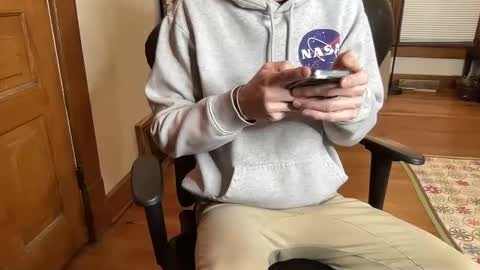 Media: A video of a person in a NASA hoodie, light pants, and black chair, holding a smartphone, in a wooden room with a door, window, and rug.