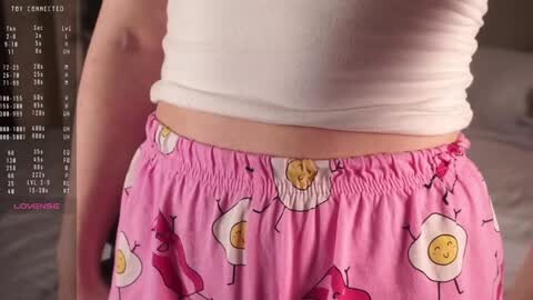Media: A close-up video of a woman's midriff in a white crop top and pink pajama pants adorned with cartoonish eggs and bananas. The background is blurred, showing a bed.