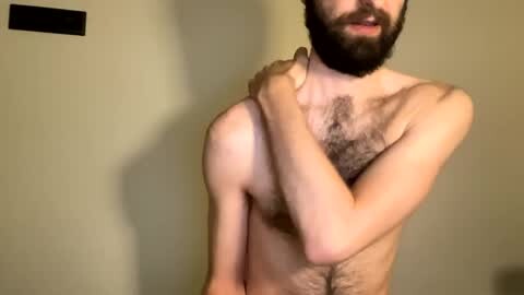 Media: Video of a bearded, light-skinned man with a hairy chest, topless, covering his chest with one arm, against a plain beige wall.
