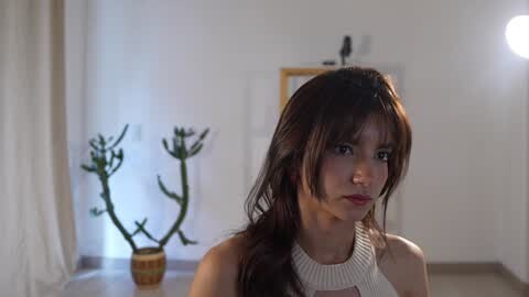 Media: Video of an Asian woman with long brown hair, wearing a white sleeveless top, in a minimalist, softly lit room with a potted cactus and a framed painting on a white wall.