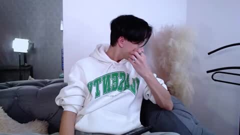 Media: Video of a young, fair-skinned, dark-haired man in a white hoodie with green text, sitting on a grey couch, covering his face, in a minimalist, modern living room with a white wall, a fluffy white pillow, and a black lamp.