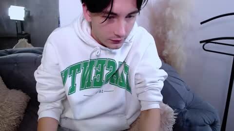 Media: Video of a young man with short dark hair, wearing a white hoodie with green \"Tigers\" logo, sitting on a gray couch in a dimly lit living room.