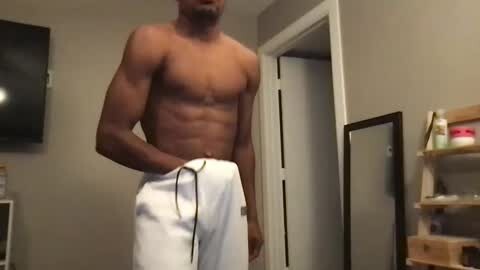 Media: Video of a shirtless, muscular Black man with dark skin wearing white athletic shorts, standing in a beige room with a flat-screen TV on the wall, shelves, and open door.