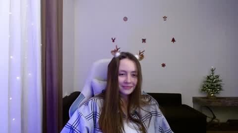 Media: Video of a young woman with long brown hair, wearing a white hat with reindeer ears, sitting in a cozy room with a Christmas tree and decorations on the wall.