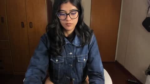 Media: Video of a young woman with medium-length, wavy black hair, wearing black-framed glasses and a denim jacket, sitting on a wooden floor in a sparsely furnished room with wooden doors.