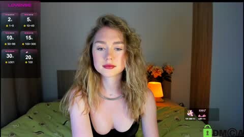 Media: A video of a blonde woman with a slender build and small breasts, wearing a black top, sitting on a bed with a green patterned cover, in a dimly lit bedroom.