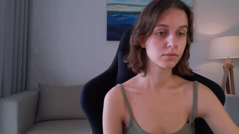 Media: Video of a young Caucasian woman with shoulder-length brown hair, wearing a light green spaghetti-strap top, sitting on a black gaming chair in a modern, softly lit room with a blue and white abstract painting on the wall.