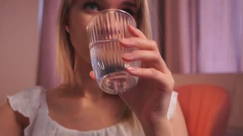 Media: Video of a young Caucasian woman with blonde hair, wearing a white sleeveless top, drinking from a glass, indoors with pink curtains and an orange cushion in the background.