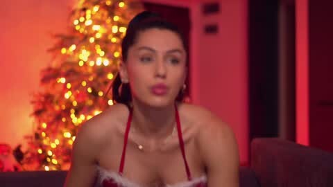 Media: Video of a light-skinned woman with dark hair in a red Santa dress, kissing a man, with a festive, warmly-lit Christmas tree in the background.