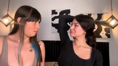 Media: Video of two young women with pale skin and dark hair, one with a black sleeveless top, the other in a black dress. They stand indoors, looking at each other. Background includes abstract art and hanging lights.