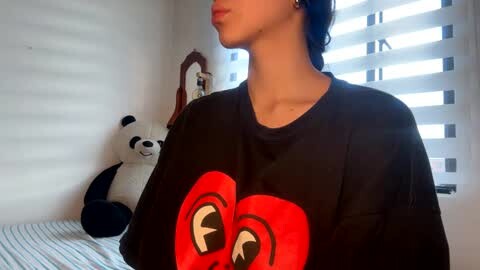 Media: Video of a woman with light skin, dark hair, and black hoodie featuring large red hearts, standing beside a window with blinds and a stuffed panda toy.