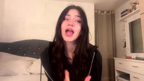 Media: Video of a young woman with long, dark hair, wearing a black hoodie, mouth open in an excited expression, standing in a cozy bedroom with a beige wall, a bed with white pillows, and a white dresser.