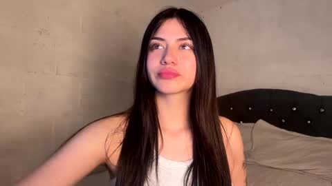 Media: Video of a young Asian woman with long black hair, wearing a white tank top, puckering her lips, standing against a beige, textured wall in a minimalist room.