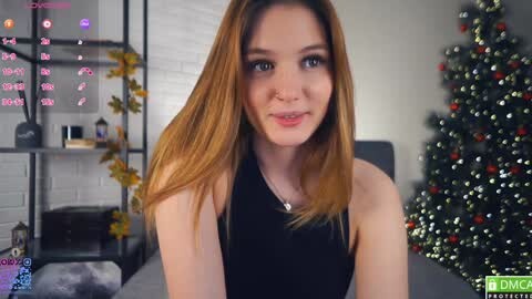 Media: A video of a young, fair-skinned woman with long, straight, blonde hair, wearing a black sleeveless top, smiling in a modern, brightly lit room with a decorated Christmas tree in the background.
