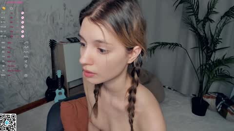 Media: A video of a young Caucasian woman with light skin and brown hair in braids, topless, sitting in a bedroom with a guitar, plants, and a potted palm tree in the background.